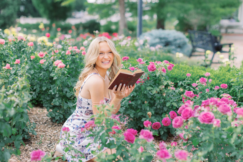 Senior girl reading a book in rose garden and laughing during her session in Lincoln